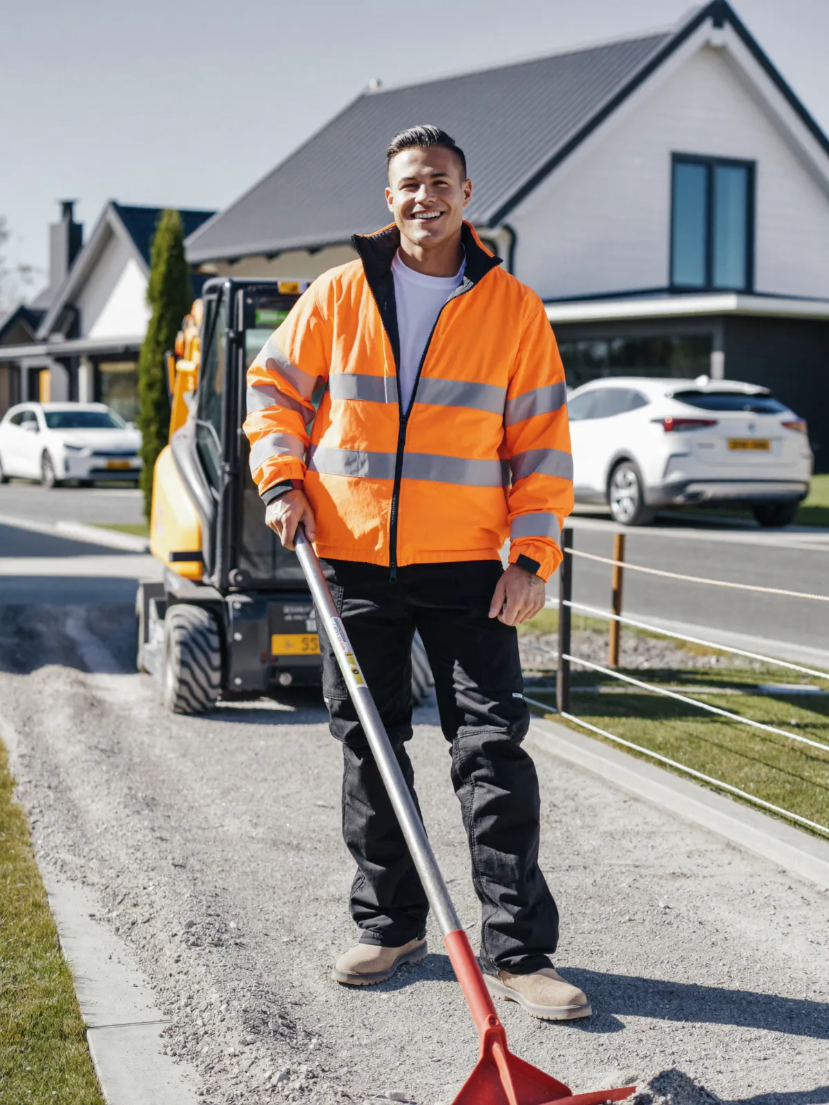A smiling groundworker in an orange hi-vis jacket with a shovel on a new gravel driveway, a mini digger behind him